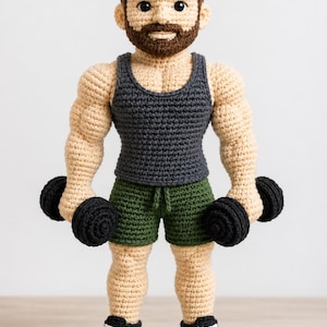 May include: A crocheted doll of a person with brown hair and a beard, wearing a dark grey tank top, olive green shorts, and black and white trainers. The doll has muscular arms and is holding a black dumbbell in each hand, standing upright.
