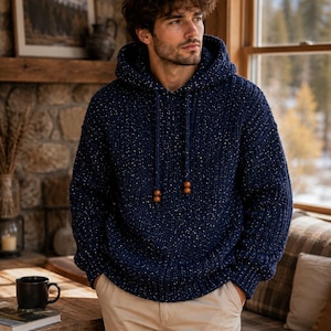 May include: A navy blue hooded sweater with white flecks and wooden toggles on the drawstring. The sweater is worn with khaki pants. The person is standing near a window with a view of a snowy landscape.