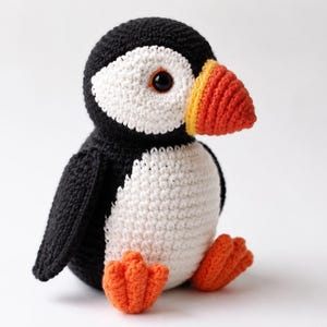 Peut inclure: Un jouet macareux en crochet fait main avec une tête et un dos noirs, un visage et un ventre blancs, ainsi qu'un bec orange vif avec une base jaune. L'animal en peluche possède deux yeux ronds sombres et deux petites pattes orange texturées.