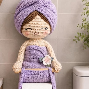 Puede incluir: Un portarrollos de papel higiénico hecho a mano con ganchillo, con forma de mujer con una toalla lavanda en la cabeza. El soporte tiene una cara sonriente, brazos y un adorno floral. El rollo de papel higiénico es visible.
