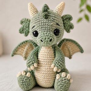May include: A handmade crochet dragon in shades of green and cream. The plush toy features large black eyes, small horns, and detailed wings. The dragon is sitting, showcasing its paws and belly.