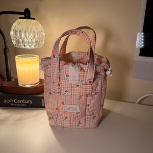 May include: A pink tote bag with a red and white checked pattern and a floral design. The bag has two handles and a white interior. The text "Love is in the air" is visible on the front of the bag.