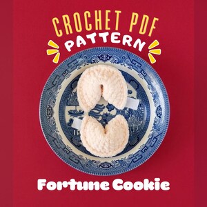 Op de afbeelding: Twee beige gehaakte gelukskoekjes rustend op een blauw-wit patroon keramieken bord. De tekst CROCHET PDF PATTERN wordt in geel en wit boven het bord weergegeven en de tekst Fortune Cookie is in het wit onder het bord gedrukt.
