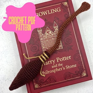 May include: A crocheted brown broom with a dark brown handle and a light brown and gold accent, resting on a red book titled "Harry Potter and the Philosopher's Stone." The image includes a pink starburst with the text "CROCHET PDF PATTERN."