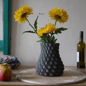 May include: A decorative gray vase with a geometric pattern holds three yellow flowers with green stems and leaves. A green wine bottle stands to the right. Two red apples and a turquoise-framed mirror are also visible.