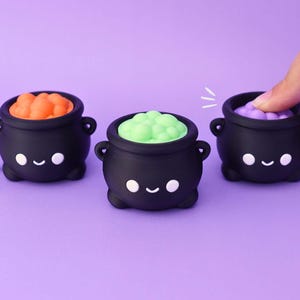 May include: Three black cauldron-shaped silicone fidget toys with smiling faces. Each cauldron features a different colored bubble top: orange, light green, and purple. One cauldron includes a metal keychain ring attached to its side.