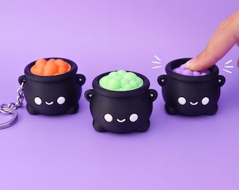 Puede incluir: Tres juguetes fidget de silicona con forma de caldero negro y caras sonrientes. Cada caldero presenta una parte superior de burbujas de distinto color: naranja, verde claro y morado. Un caldero incluye una anilla metálica para llaves.