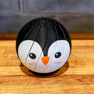 May include: A 3D-printed spherical puzzle featuring a penguin design. The penguin has a black body, white face, and an orange beak. The puzzle is made of interlocking pieces and sits on a wooden surface.