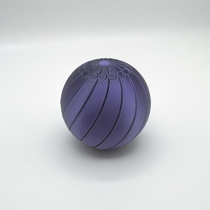 Op de afbeelding: Een bolvormig object in paars, met een geribbeld ontwerp en gebogen lijnen. De bovenkant van de bol heeft een gedetailleerd, patroonontwerp. Het object staat tegen een effen witte achtergrond.