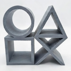 May include: A gray, 3D-printed sculpture featuring a circle, square, triangle, and an X-shaped diamond. The geometric shapes are arranged together, creating a modern art piece. The sculpture is made of a solid, matte material.