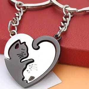 May include: Two-piece cat-shaped keychains. Each keychain forms half of a heart when joined. One cat is black, the other is silver. The keychains are connected to a silver chain and ring. The background is a red box.