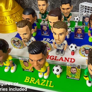 May include: A collection of football bobbleheads, featuring player likenesses. The figures are displayed on green and blue bases, with the words "BRAZIL" and "ENGLAND". Small footballs and player cards are also visible. A golden FIFA World Cup trophy is in the background.