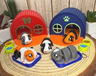 May include: A collection of colorful guinea pig figurines and miniature houses. The houses are red and blue with orange bases. The guinea pigs are in various poses, some in their houses, others on blue trays. There are also small food bowls.