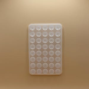 Puede incluir: Un molde de silicona transparente y rectangular con 42 cavidades redondeadas y espaciadas uniformemente. El molde está diseñado para crear pequeños objetos esféricos. El fondo es de color marrón claro liso.