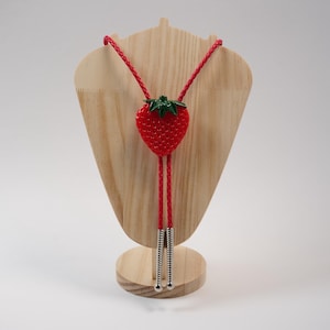 May include: A bolo tie featuring a bright red strawberry pendant with a green stem. The cord is made of braided red material, and the two ends are finished with silver-toned metal tips. The strawberry has a textured surface with small dots.