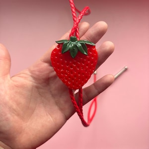 Op de afbeelding: Een rode aardbeivormige bolo-stropdas met groene bladeren en een gevlochten rood koord. De bolo-stropdas heeft een zilverkleurige metalen sluiting. De aardbei is felrood.