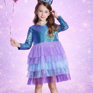 Może przedstawiać: Niebiesko-fioletowa sukienka o tematyce syreny z długimi rękawami i warstwową tiulową spódnicą. Sukienka ma wzór łusek na gorsecie i jest połączona z opaską na głowę z rogiem jednorożca. Różowy balon trzymany jest w lewej ręce.