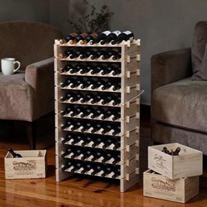 May include: A tall, light-colored wooden wine rack filled with wine bottles. The rack has multiple shelves and is surrounded by two wooden crates. The wine rack is in a living room setting with a couch and chair.