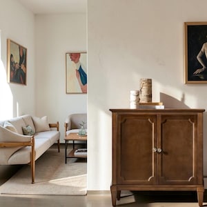 May include: A medium-brown wooden cabinet with two doors, featuring recessed panels and small round silver knobs. The cabinet stands on four tapered legs and is topped with two cylindrical birch bark decorative accents and a small book.