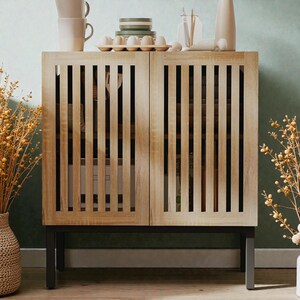 May include: A light wood cabinet with two doors featuring vertical slat detailing. The cabinet sits on a black metal frame. The top of the cabinet displays white mugs, plates, and eggs. Dried floral arrangements flank the cabinet.