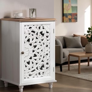 May include: A white cabinet with a natural wood-toned top surface. The single door features an intricate, carved floral mandala pattern and a small round knob. The cabinet is supported by four short, turned feet and has a rectangular frame.