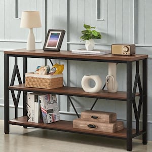 May include: A three-tiered, dark brown and black console table with decorative items. The top shelf holds a lamp, framed picture, and a small radio. The middle shelf has a woven basket and white vases. The bottom shelf has decorative boxes and books.