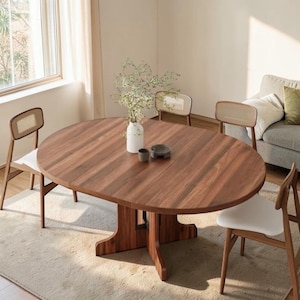 May include: A large, oval wooden dining table with a dark brown finish. The table is supported by a pedestal base and surrounded by four chairs with woven backs and white seats. A vase of greenery and decorative bowls sit on the table.