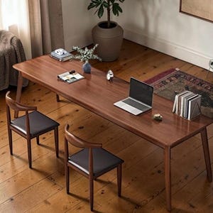 May include: A long, rectangular wooden table with a dark finish, accompanied by two chairs with dark seats. A laptop, books, and decorative items are arranged on the table. The scene is set on a hardwood floor, with a rug visible in the background.
