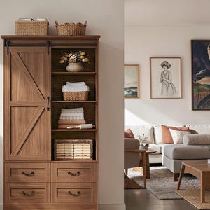 May include: A tall brown wooden storage cabinet featuring a sliding barn-style door with a diagonal cross brace, four lower drawers with dark metal pulls, and open shelving containing woven baskets, folded linens, and a decorative vase.
