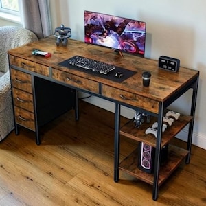 May include: A brown and black gaming desk with a monitor, keyboard, and computer tower. The desk has a wood grain finish and a black metal frame. The desk has drawers on the left side and shelves on the right side. A small clock displays the time.