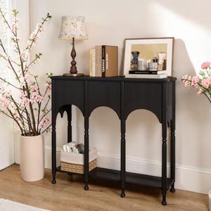 May include: A black console table with arched supports and a lower shelf. A lamp, books, and a framed picture sit on top. A vase of pink flowers and a basket are also visible.