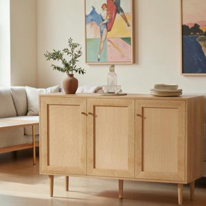 May include: A light wood sideboard featuring three cabinet doors with woven cane panels and small round knobs. The piece stands on four tapered legs and is topped with a terracotta vase containing green branches, a glass bottle, and stacked bowls.