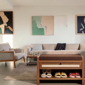 May include: A wooden shoe storage bench with a brown padded seat and a single drawer, featuring open shelving below containing four pairs of sandals. The bench is positioned in a living room with a beige sofa, an armchair, and framed wall art.