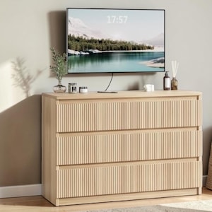 May include: A light wood-tone dresser with three drawers, each featuring a vertical grooved design. A flat-screen TV displaying a landscape scene is mounted above. The time 17:57 is displayed on the TV screen. Decorative items sit on top of the dresser.