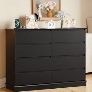 May include: A black dresser with eight drawers, featuring a wood grain texture. The dresser has a rectangular shape and is topped with decorative items, including a phone charger, a book, and small figurines. The overall design is modern and functional.