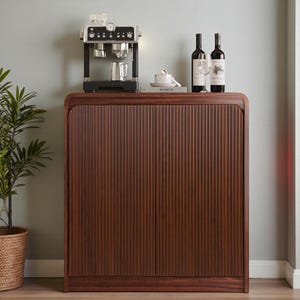 May include: A dark brown wooden cabinet with vertical grooved detailing. A coffee machine, wine bottles, and a cup and saucer are on top. A potted plant is on the left.