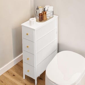 May include: A white, narrow bathroom storage cabinet with four drawers and gold-colored knobs. A wire basket on top holds toiletries, and a roll of toilet paper sits on the cabinet's surface. The cabinet is positioned next to a white toilet.