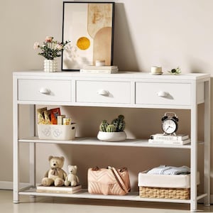 May include: A white metal console table with three drawers and two shelves. The top shelf holds a vase of flowers, books, and a framed abstract art print. The lower shelves display a variety of decorative items, including a teddy bear, cactus, and a purse.