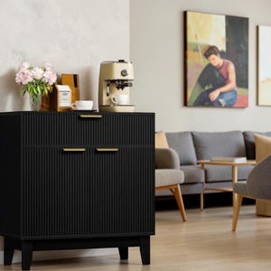 May include: A black cabinet with a vertical ribbed texture and gold-toned horizontal handles. The cabinet features a top drawer and two lower doors, supported by four short legs. A beige espresso machine and a glass vase with pink flowers sit on top.