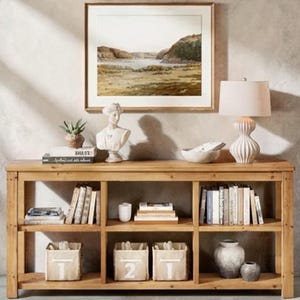 May include: A wooden console table with open shelving, displaying books, decorative baskets, and a bust. A framed landscape painting hangs above, with a lamp and a bowl on the tabletop. The overall aesthetic is rustic and elegant.