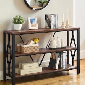 May include: A three-tiered, dark brown and black console table. The table has a rectangular top and shelves with a black metal frame. Decorative items include books, candles, and a small potted plant. The table is 48 inches long.