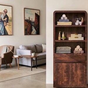May include: A tall, dark wood shelving unit with four open shelves and a bottom cabinet. The shelves hold stacks of folded towels, small bottles, and two rectangular boxes. The unit stands next to a living room with a sofa, chair, and wall art.