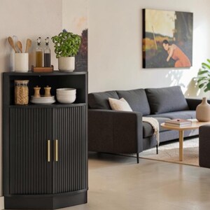 May include: A black corner cabinet with vertical fluted doors and gold handles. The top surface holds a white ribbed utensil holder, two glass oil bottles, and a potted green plant. A glass jar with contents, small wooden items, and white bowls are also present.
