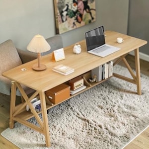 May include: A light-colored wooden desk with a laptop, lamp, books, and a small alarm clock. The desk has a lower shelf with storage boxes and books. The desk is set on a fluffy, light-colored rug. The desk is approximately 60 inches long.