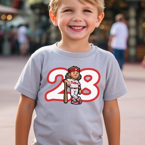 May include: A light gray t-shirt featuring a cartoon baseball player with the number 28 in a red outline. The player is wearing a red headband and holding a baseball bat. The shirt is worn by a smiling child.