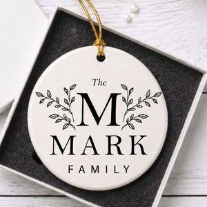 May include: A white ceramic ornament, 3 inches in diameter, with a gold hanging string, displayed in a gift box. The ornament features the text "The MARK FAMILY" with a large initial "M" and leaf accents.