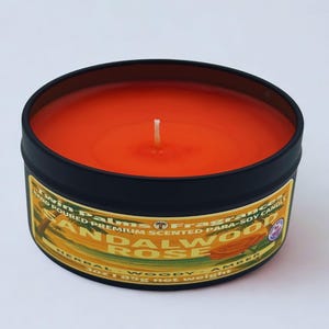 A round, black tin candle with an orange wax interior and a white wick. The label reads "Twin Palms Fragrances Sandalwood Rose" with "Herbal Woody Amber" below. The candle weighs 3 oz (85g).