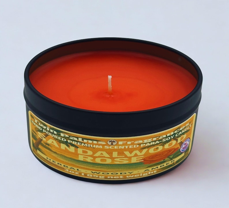 A round, black tin candle with an orange wax interior and a white wick. The label reads "Twin Palms Fragrances Sandalwood Rose" with "Herbal Woody Amber" below. The candle weighs 3 oz (85g).