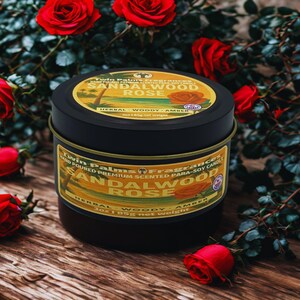 A black candle tin with a gold label that reads "Sandalwood Rose." The candle is surrounded by red roses on a wooden surface. The label also states "Herbal, Woody, Amber" and "3oz 85g net weight."