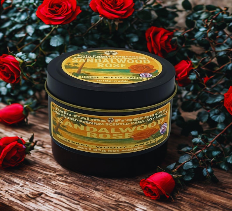 A black candle tin with a gold label that reads "Sandalwood Rose." The candle is surrounded by red roses on a wooden surface. The label also states "Herbal, Woody, Amber" and "3oz 85g net weight."